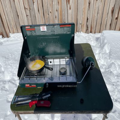 A picture of a gas propane cooking eggs outside in the snow.